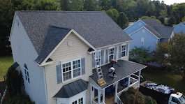 Workers on a two-story house roof, one on a ladder. Blue sky, beige siding, black roof.
