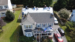 Roofing workers on a house roof. Blue siding, green grass, and vehicles in the driveway.