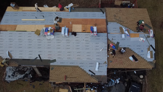 Overhead view of roofers working on a house roof replacement; shingles, plywood, and materials are visible.