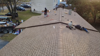 Roofers installing shingles on a residential roof in progress. Brown roof, workers, tools, and a trailer with debris.