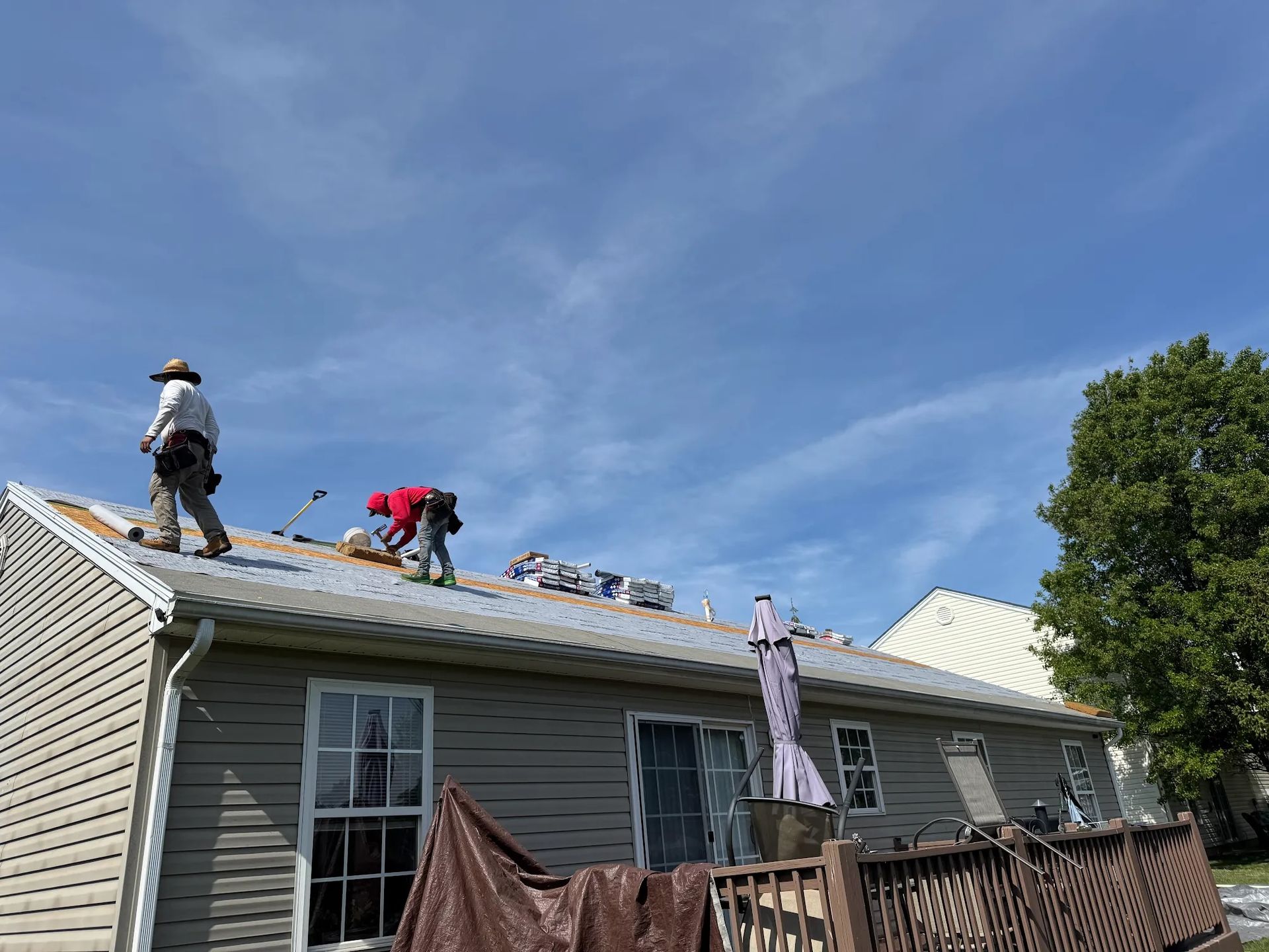 Roofers working on a house roof under a blue sky, removing old shingles and installing new ones.