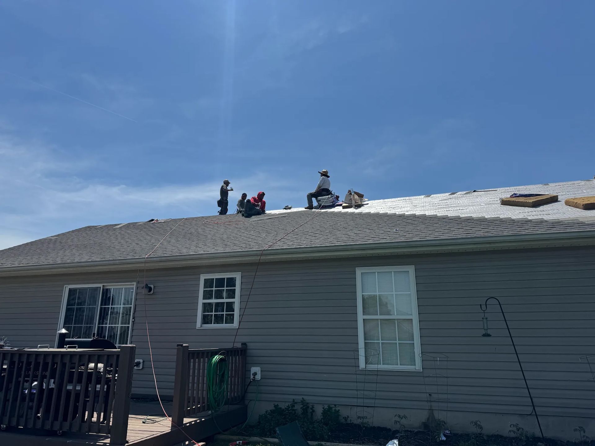 Roofers on a gray shingled roof under a bright blue sky. A gray house with white windows is visible.