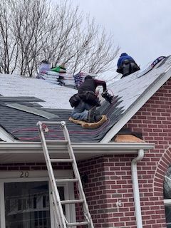 Roofers replacing shingles on a brick house. Ladder propped against the roof. Bags of shingles visible.