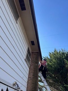 Person on a ladder, near a brick chimney, works on a building's gutter under a blue sky.