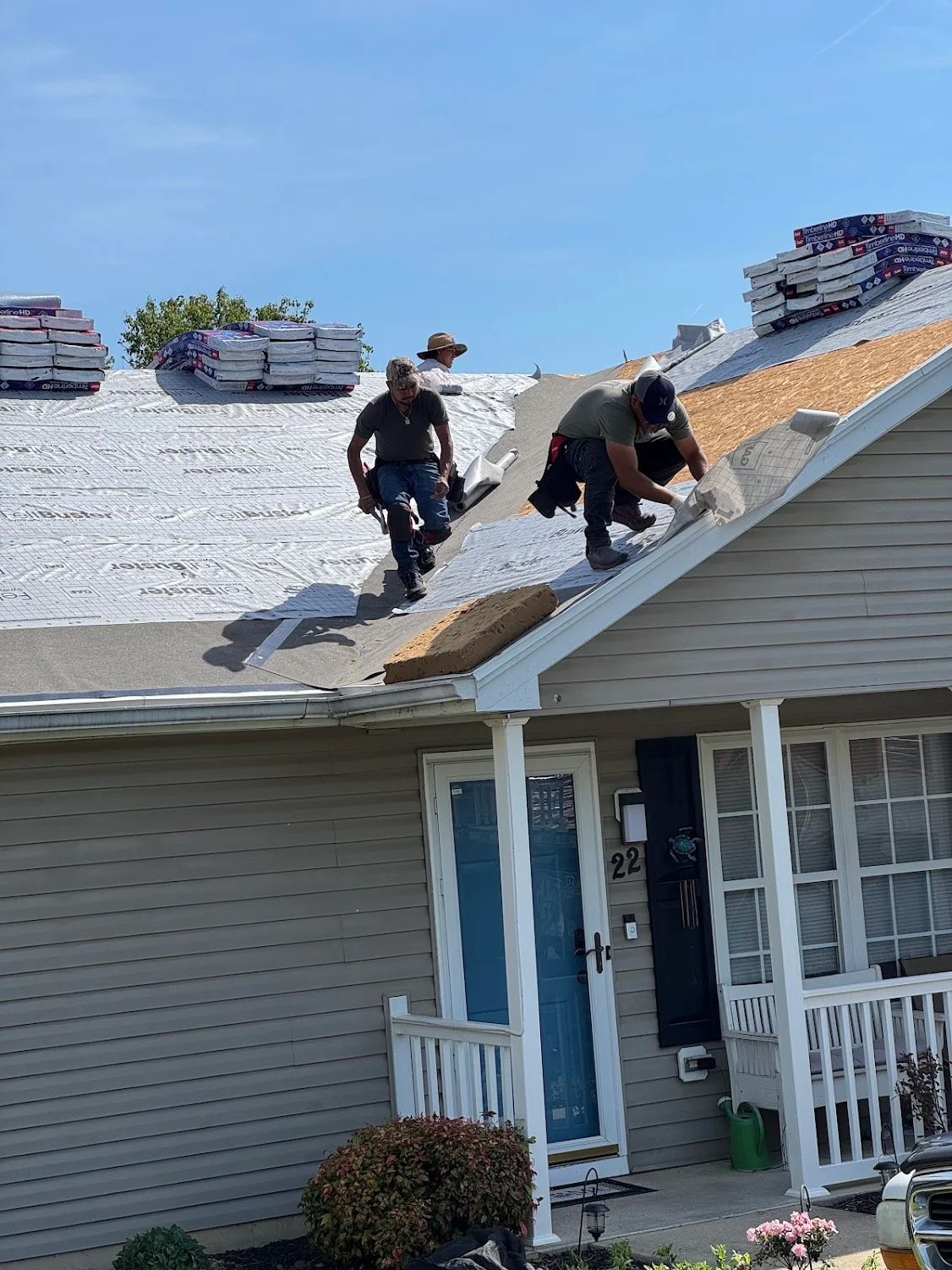 Roofers working on a house, with stacks of shingles on the roof under a blue sky.