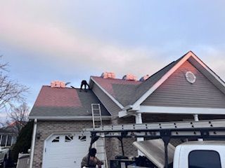 Workers on a roof with stacks of shingles, a ladder, and a truck below. Gray shingles, cloudy sky.