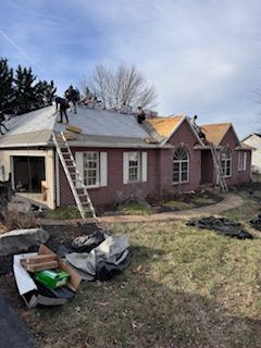 Roofers working on a red brick house; ladders, materials, and debris visible on the ground.
