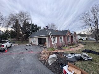Workers on a roof of a brick house with a garage; a ladder is propped against the roof. Cloudy sky.