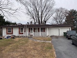 A white ranch-style house with roofing work in progress; ladder, tools, and parked vehicles visible.