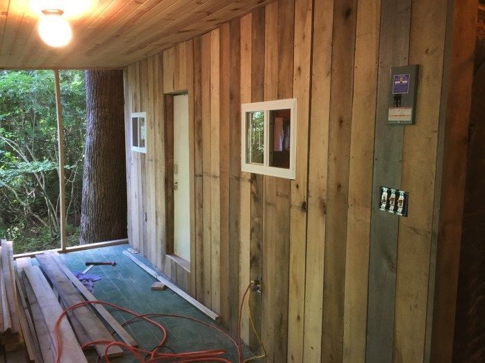 Wooden-walled room under construction, with small windows and door.  Tools and materials on the floor, and a large tree trunk visible outside.