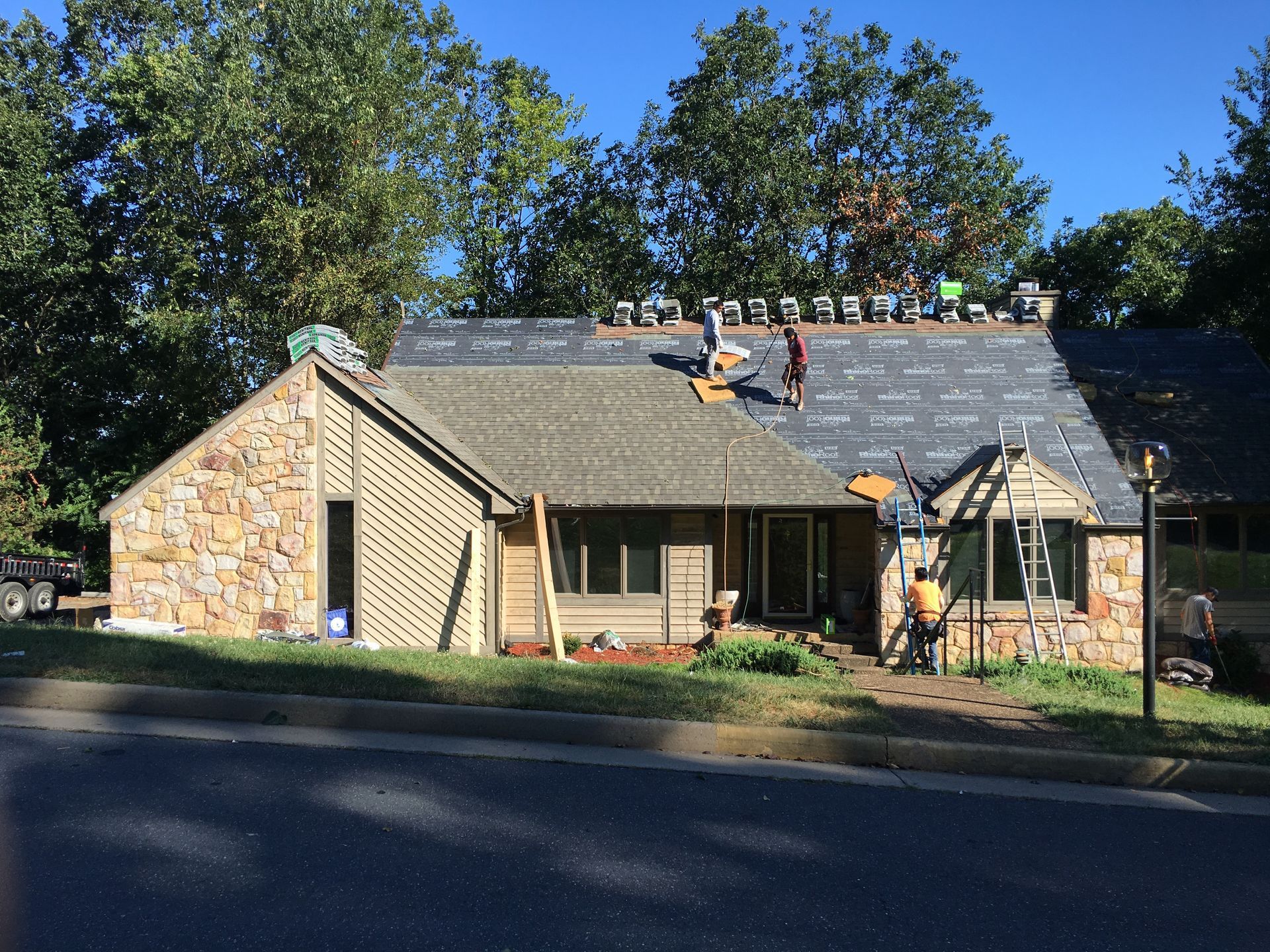 Workers replacing a roof on a stone-faced house. Blue sky, tools, and materials are visible.