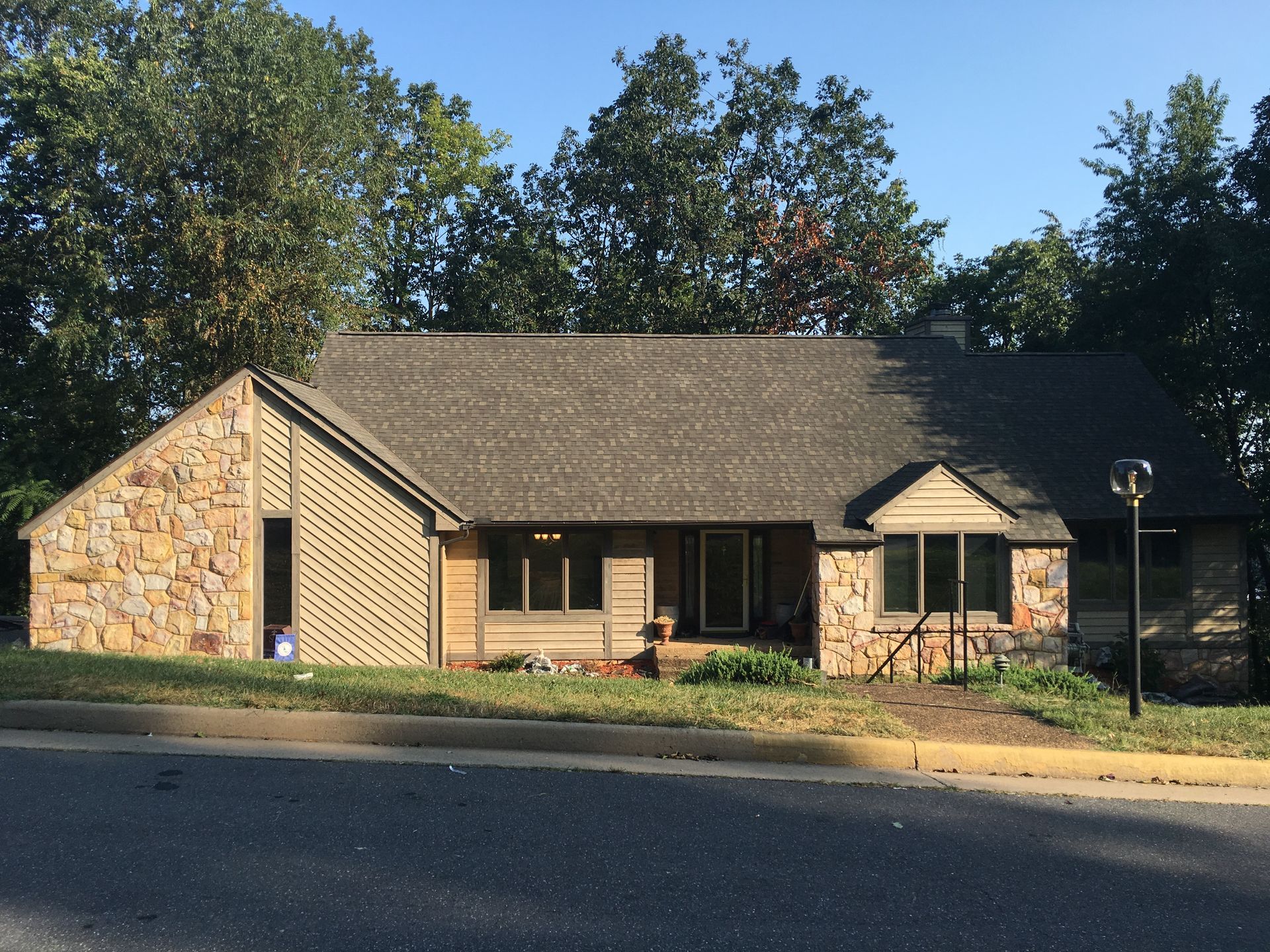 A single-story house with stone and wood siding, a gray roof, and trees in the background.