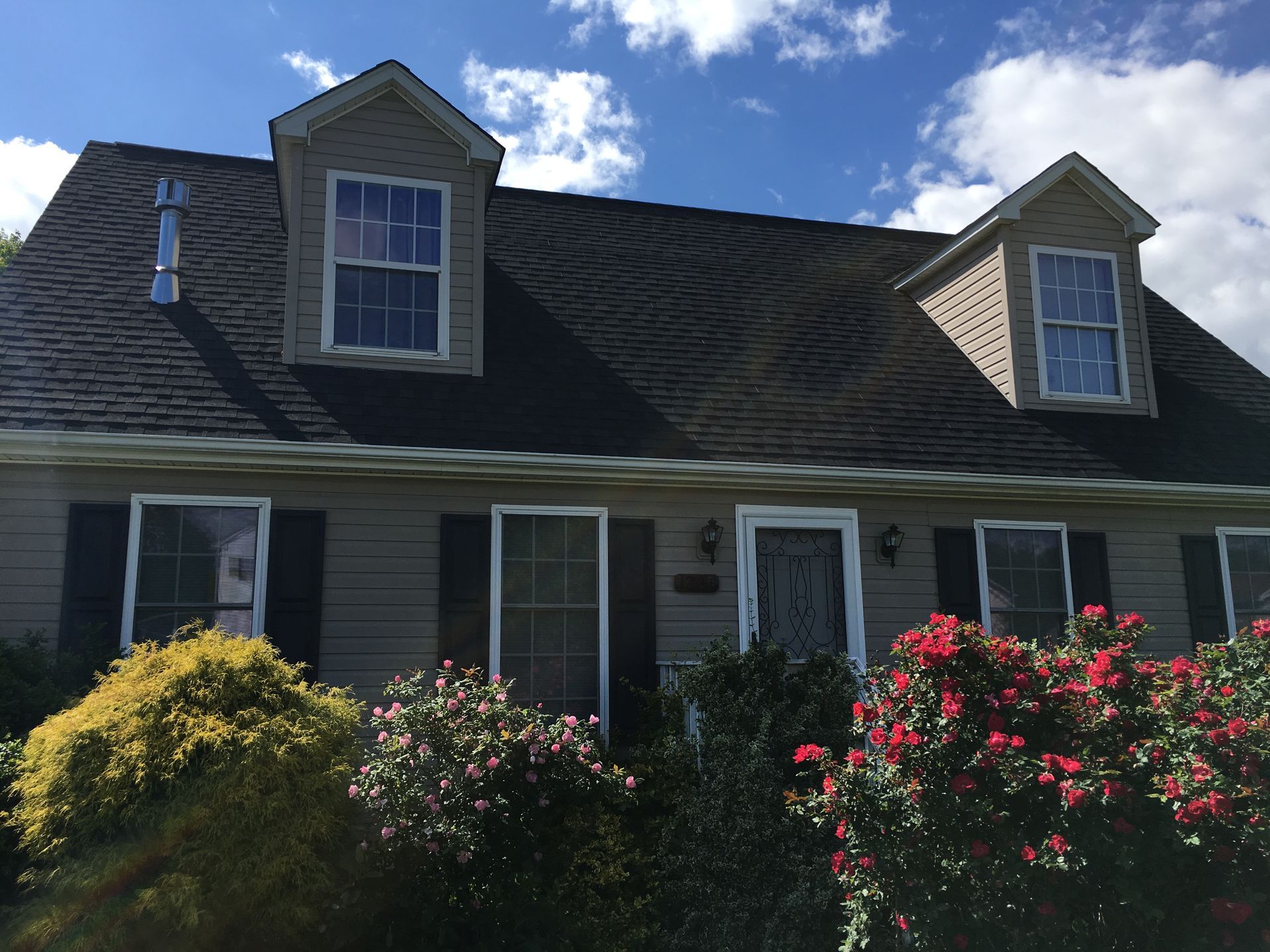 Tan house with black roof, two dormers, and bushes in front. Blue sky with clouds.