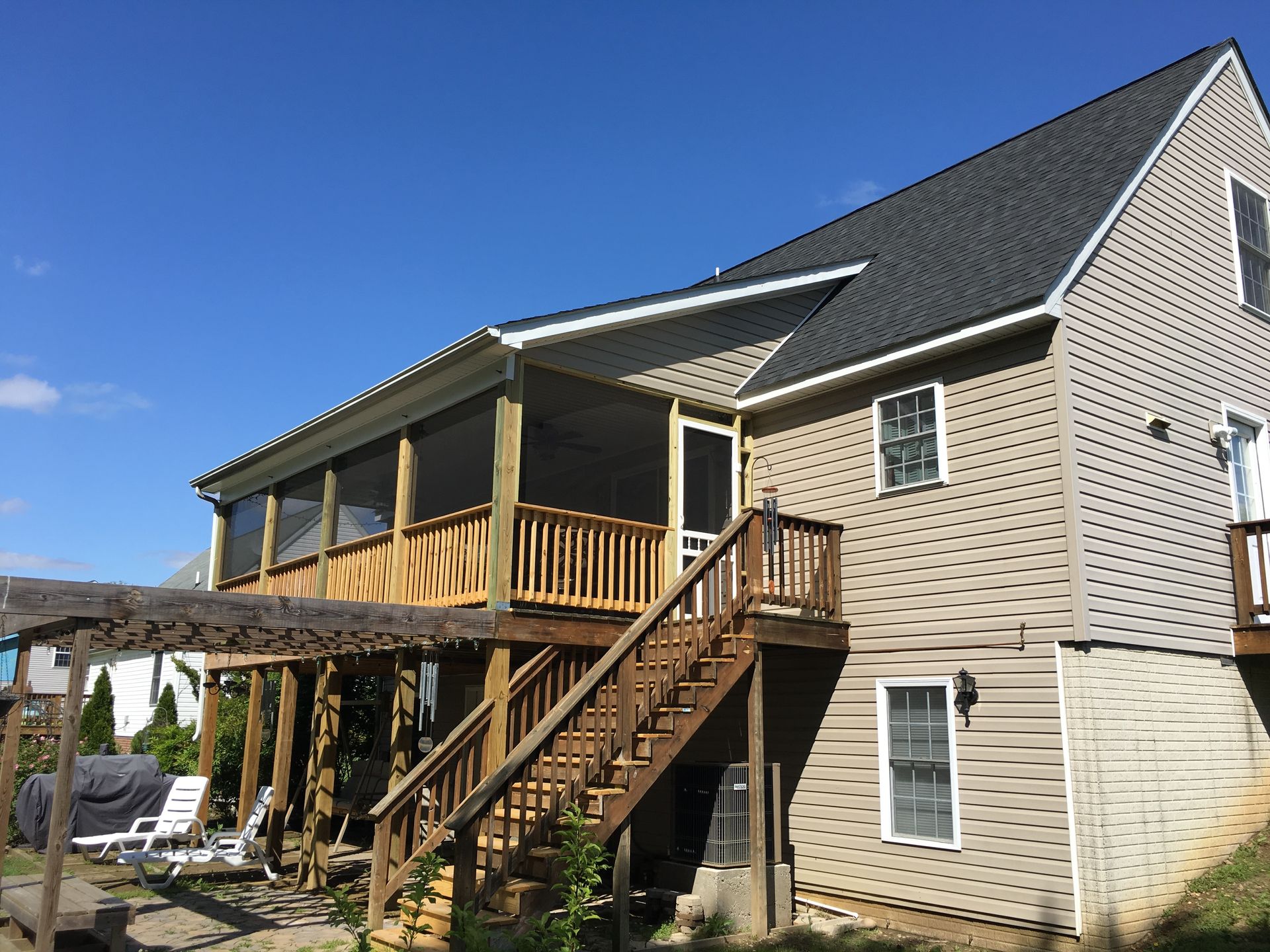 Back of a two-story house with a wooden deck, screened porch, and stairs against a clear blue sky.