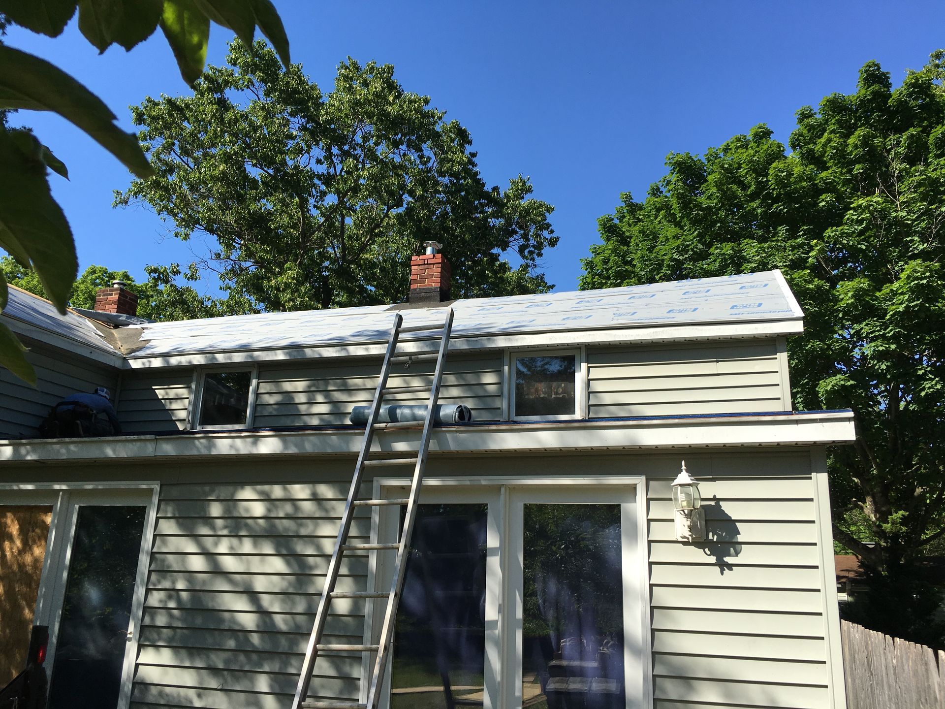 Ladder leaning against a house with a gray roof and chimneys; trees and blue sky background.