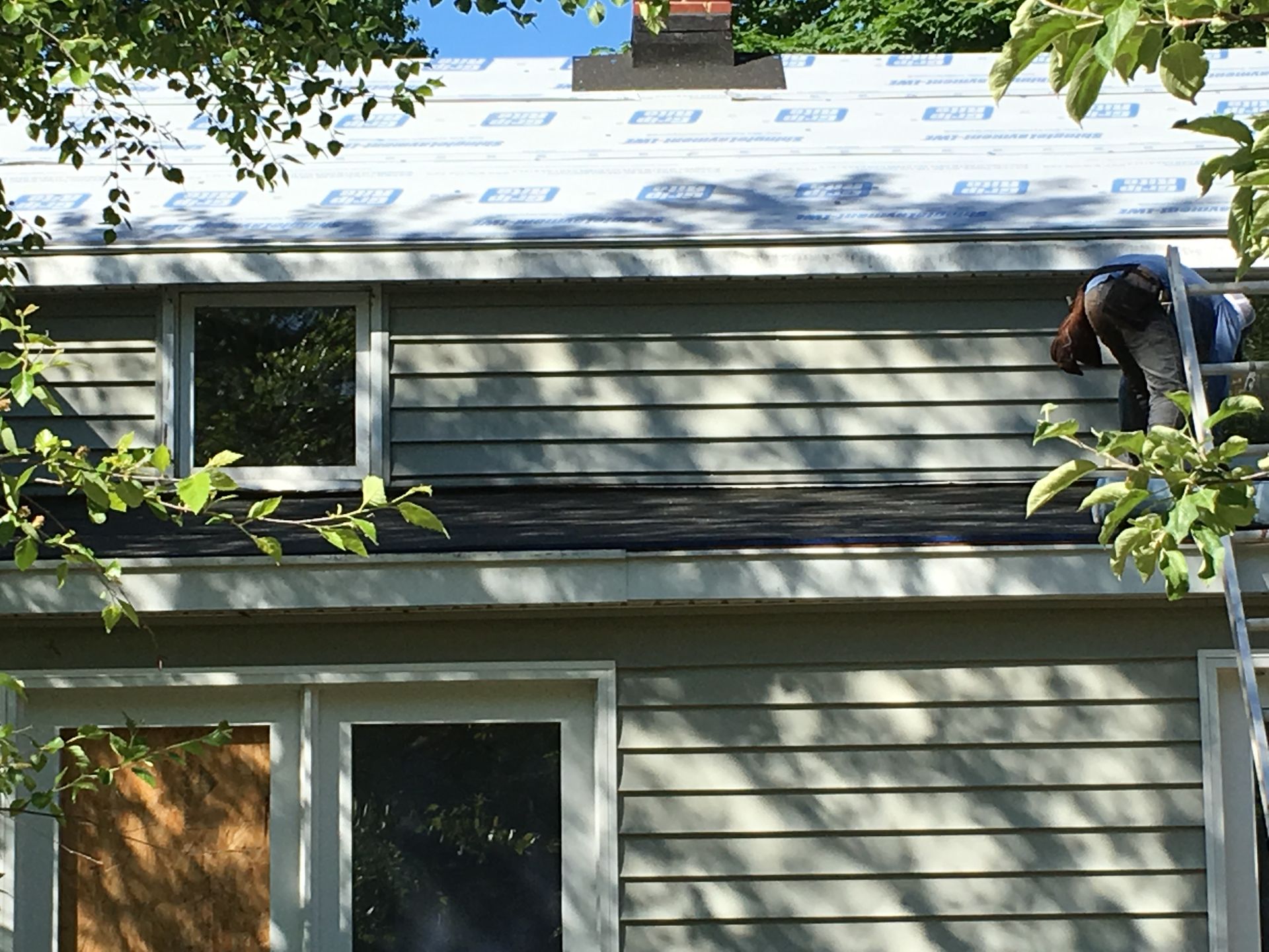 Man on roof installing roofing material; house with green siding, windows, and trees.