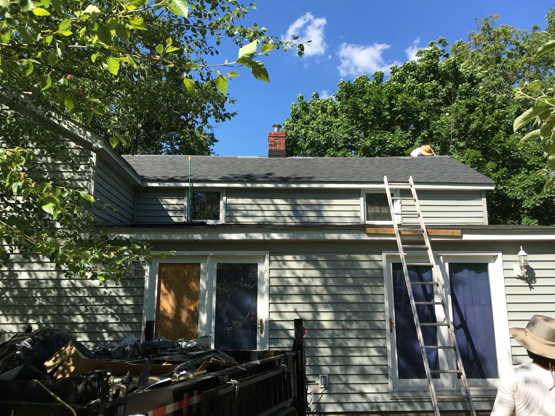 House roof repair; ladder propped up; trash bin below; surrounded by trees and blue sky.