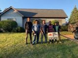 Five people stand in front of a house, holding a sign. Sunny day, green grass, light blue house.