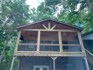 Wooden cabin with porch, railing, and triangular roof design, surrounded by trees.
