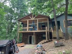 House under construction with a deck, surrounded by trees. Blue house. Construction materials visible.