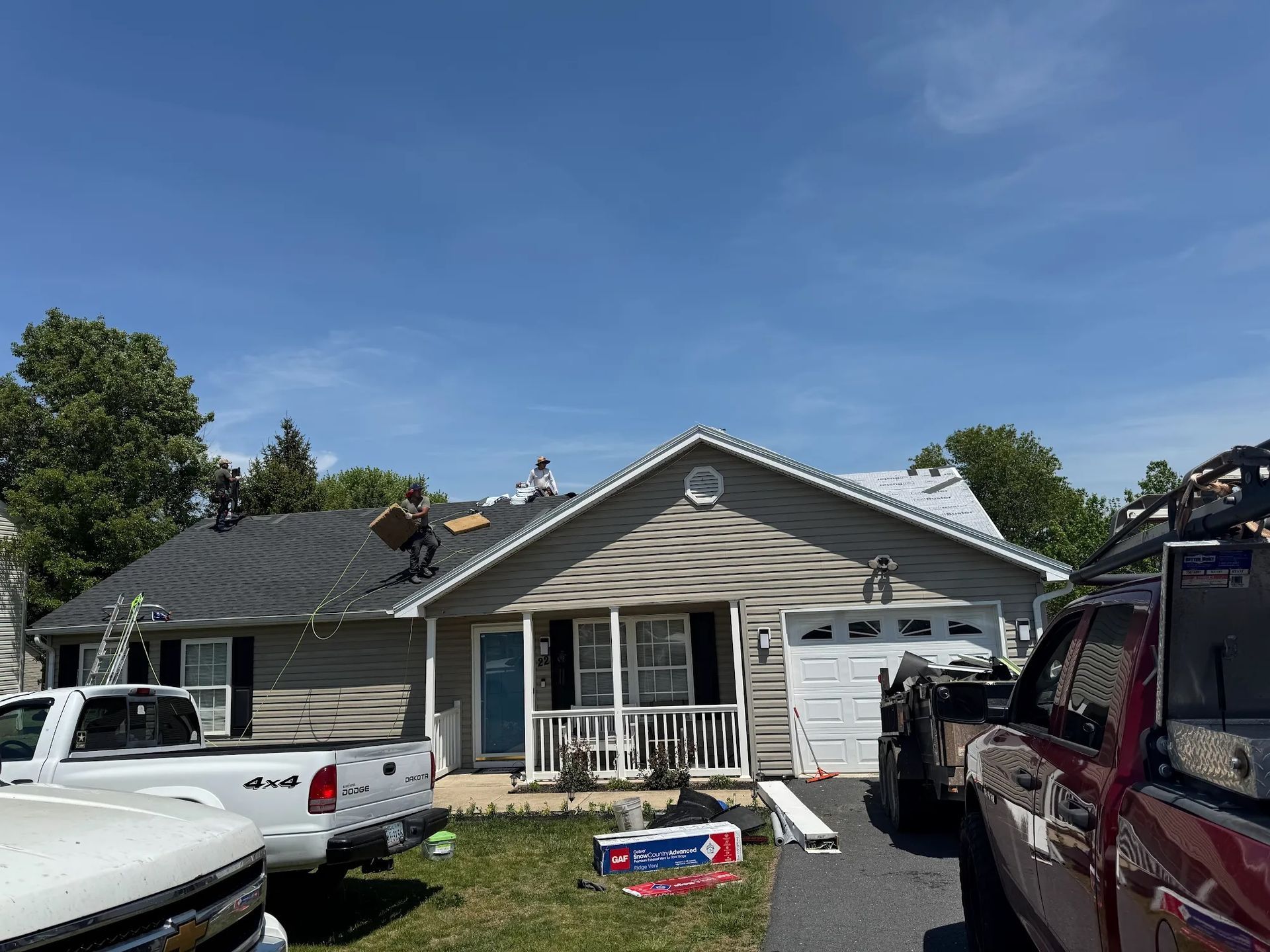 Roofers working on a residential home with gray siding. Trucks parked in the driveway on a sunny day.