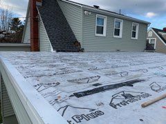 Flat roof covered with black membrane, attached to a light green house with multiple windows.
