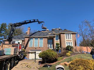 Crane lifting roofing materials onto a two-story brick house with workers on the roof under a clear blue sky.