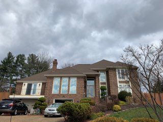 Large brick house with brown roof and cloudy sky. Cars in the driveway, landscaping in front.