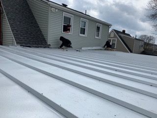 Two workers on a metal roof of a light-colored building, installing or repairing it. Overcast sky.