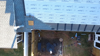 Overhead view of a house roof partially covered with shingles, with construction tools and materials in the yard below.