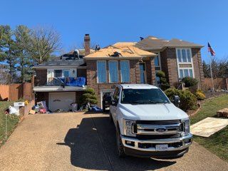 White truck parked in front of a house with roofers working on the roof.  Blue tarp covers a section. Sunny day.