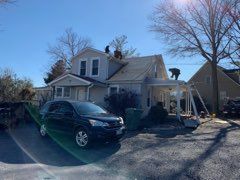 Workers on a house roof replacing shingles. Black car in driveway. Sunny, daytime.