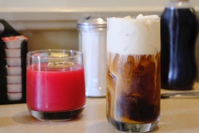 Tall glass of iced coffee topped with cream next to a small glass of red liquid; sugar dispenser and syrup bottle in background.