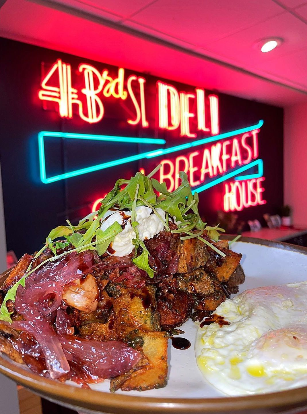 Dish with potatoes, onions, greens, and fried egg. Neon sign for a breakfast deli in background.
