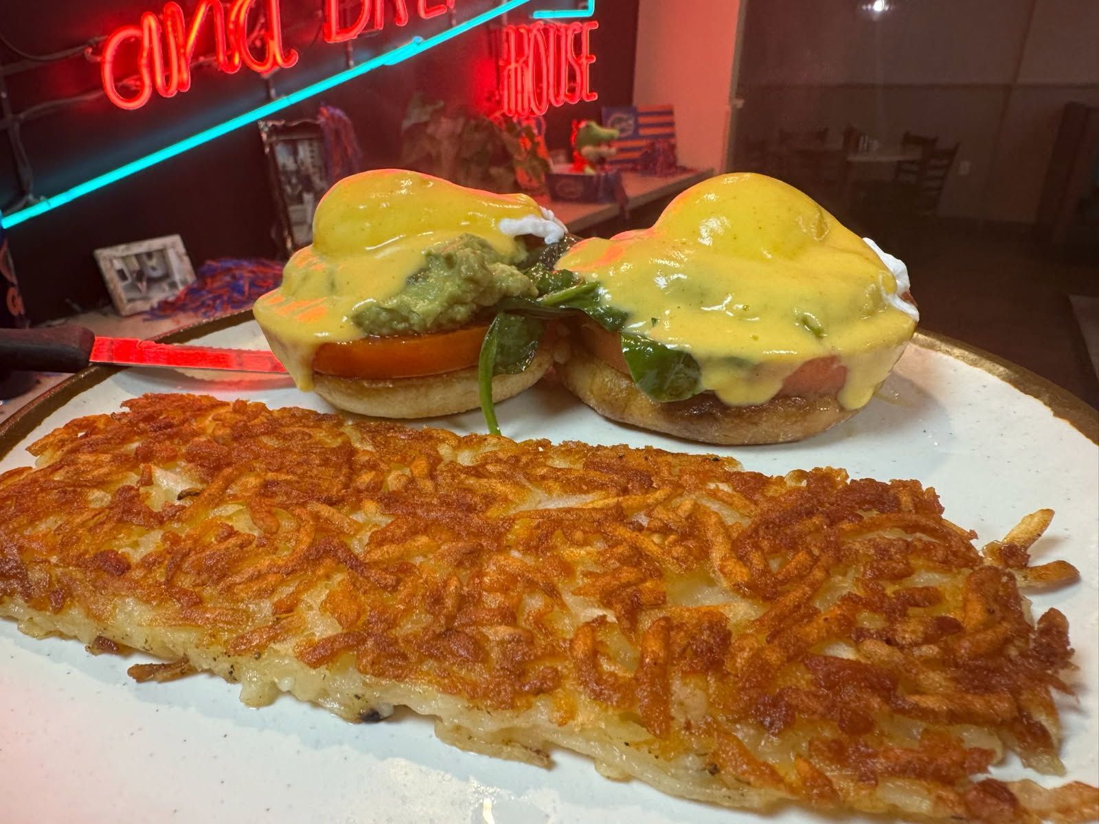 Eggs Benedict with hash browns on a plate. Neon sign in background.