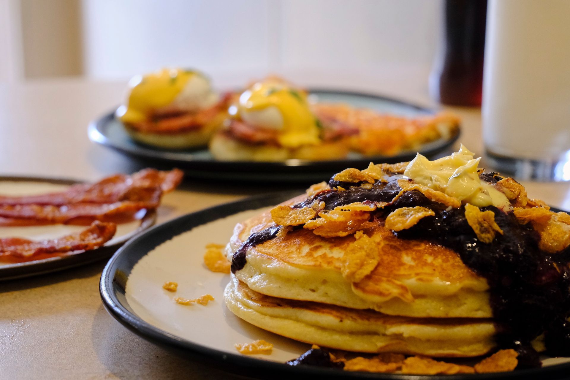 Stack of pancakes with syrup and corn flakes, bacon, and Eggs Benedict on a table.