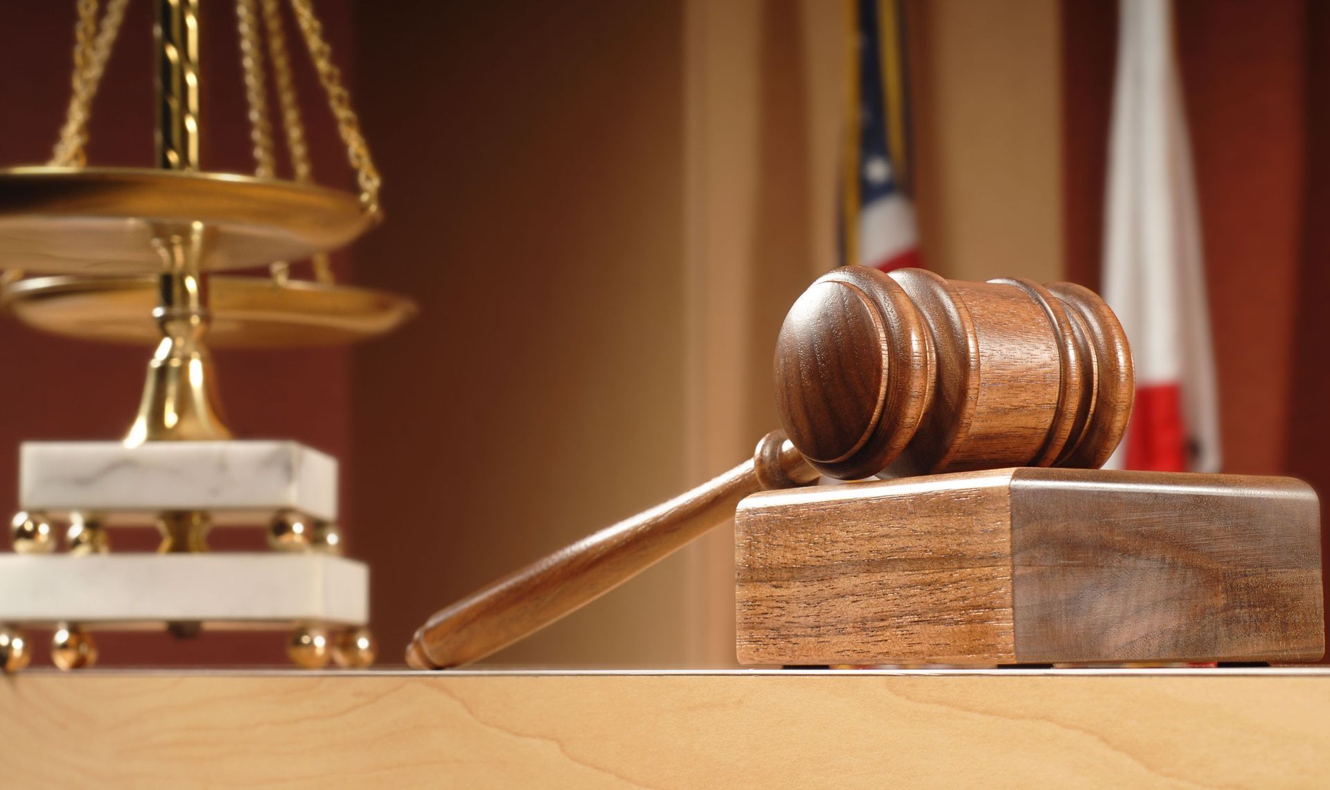Wooden judge’s gavel on a block beside a balanced scale in a courtroom setting.