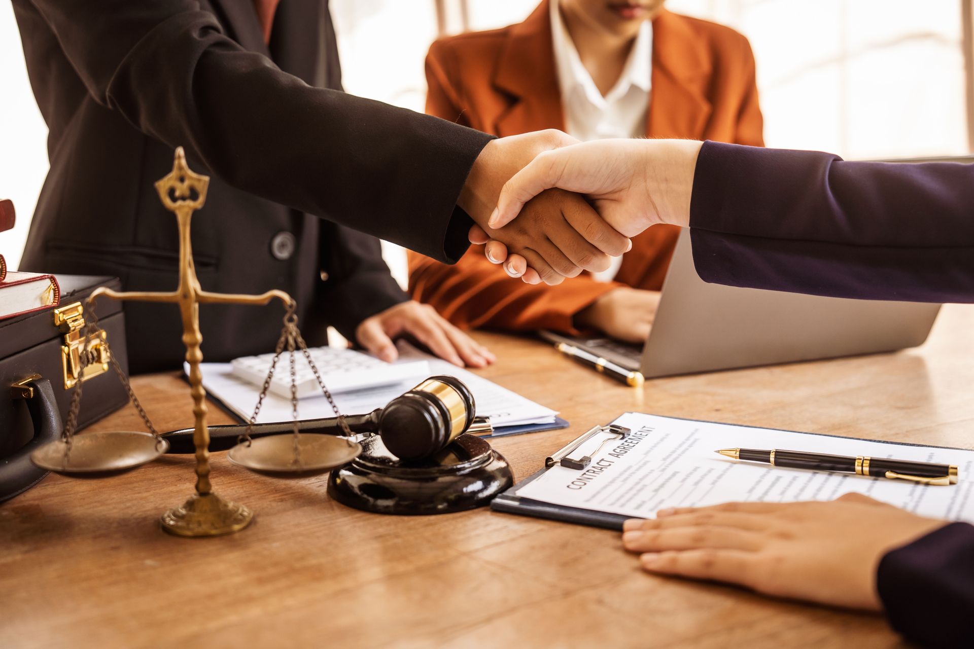 Lawyers shaking hands over a desk with a scale, gavel, and documents in a courtroom office