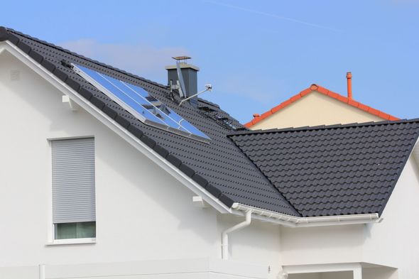 White house with black tiled roof, solar panels, and a window with a closed blind under a blue sky.