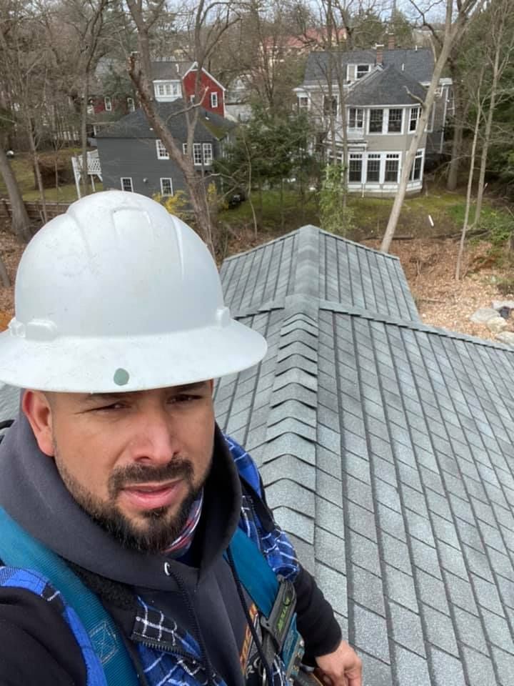 Man in white hard hat on a gray shingled roof, looking at the camera. Buildings and trees in the background.
