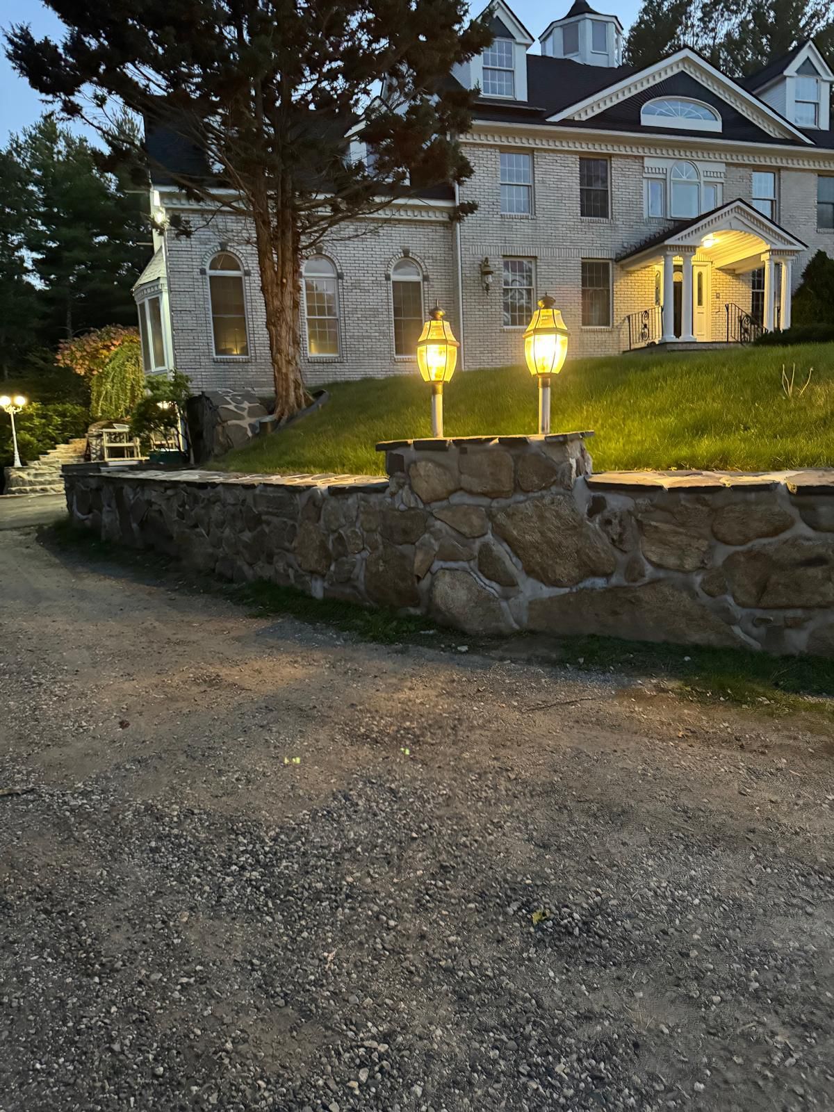 Gravel driveway leading to a large stone house at dusk. Lanterns on stone wall illuminate the entrance.