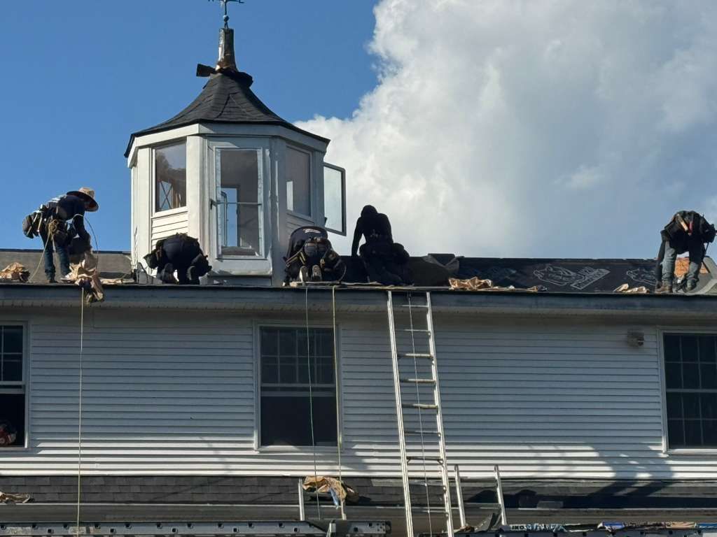 Roofers working on a white house roof with a cupola and ladder, blue sky in background.