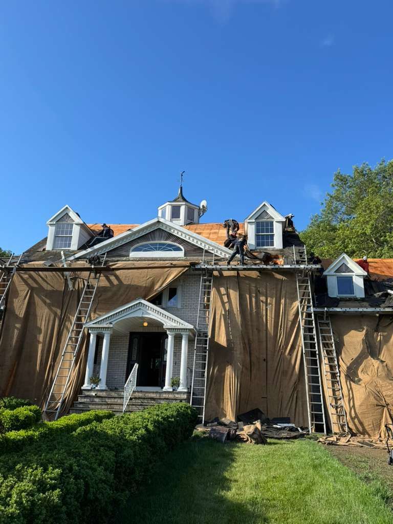 Workers on roof of house under construction; brown tarp covers facade. Blue sky.