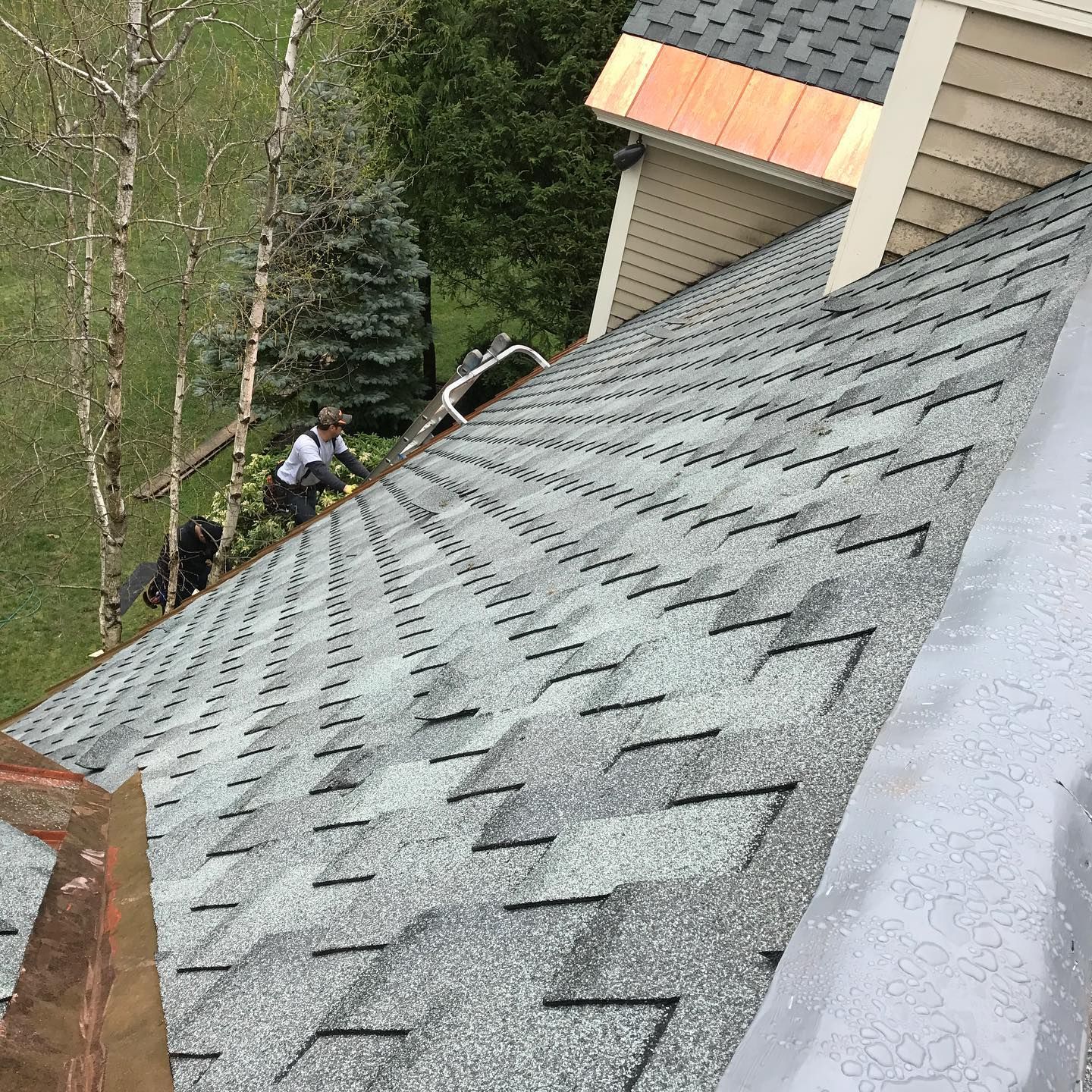 Person working on a gray shingle roof, green grass and trees in the background, angled view from above.