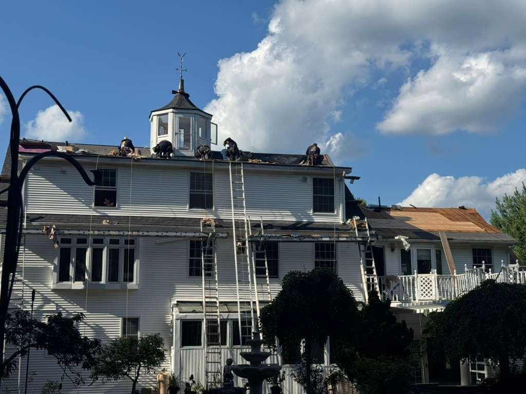 Workers on a white building roof, ladders against the side. Blue sky, partly cloudy.