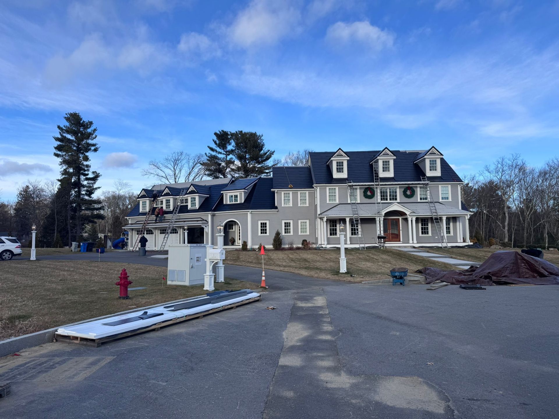 Large gray house with black roof, dormers, and Christmas wreaths. Outdoor setting with cloudy sky.
