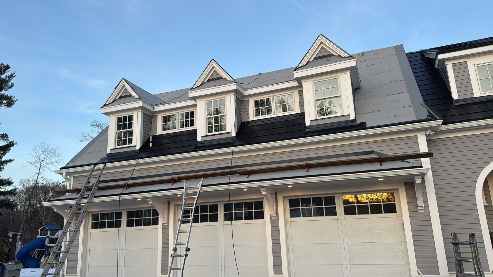 Garage with gray siding, white trim, dormers with windows, and ladders.