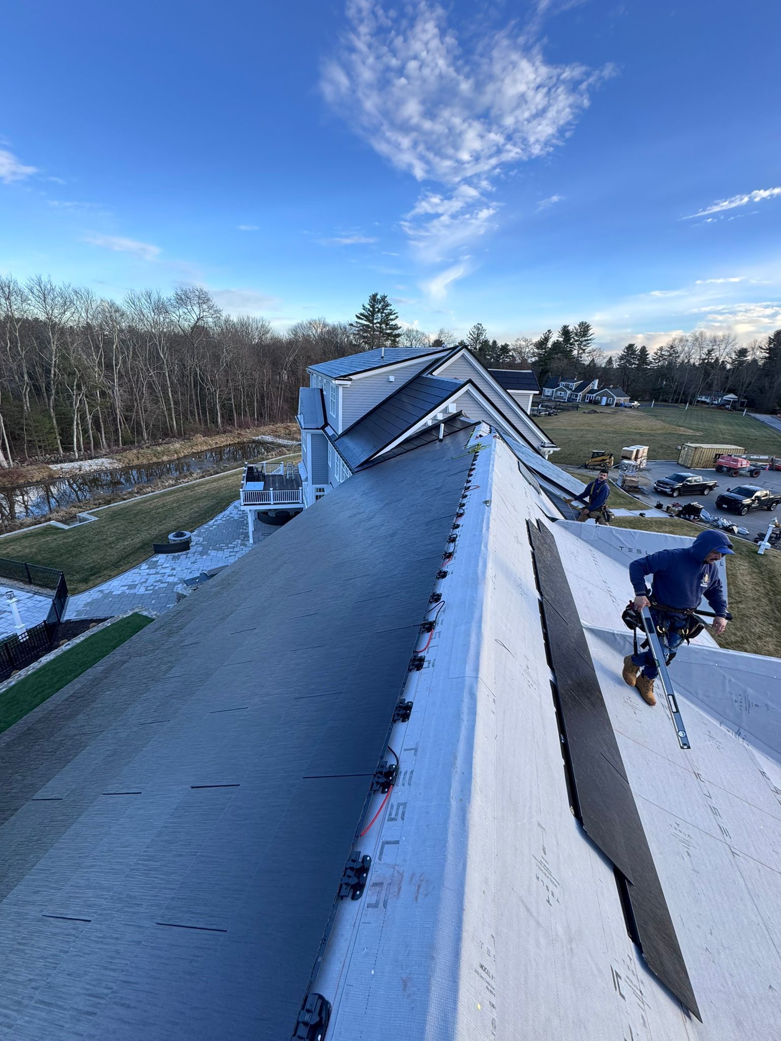 Roofer working on a dark roof with other workers in the distance under a blue sky.