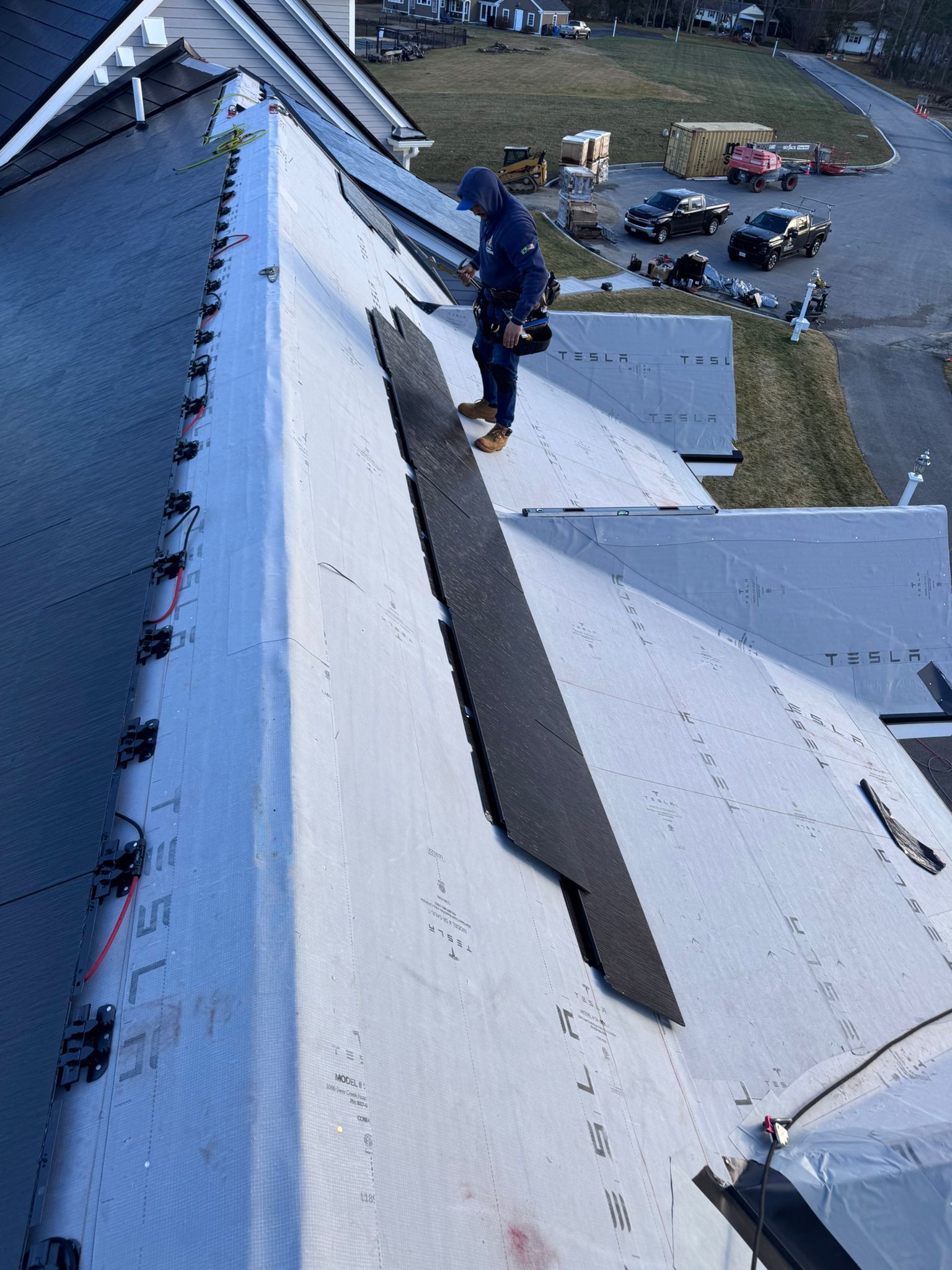 Roofer on a light-colored roof, installing a black ridge cap. Blue workwear, clear daylight, residential area.