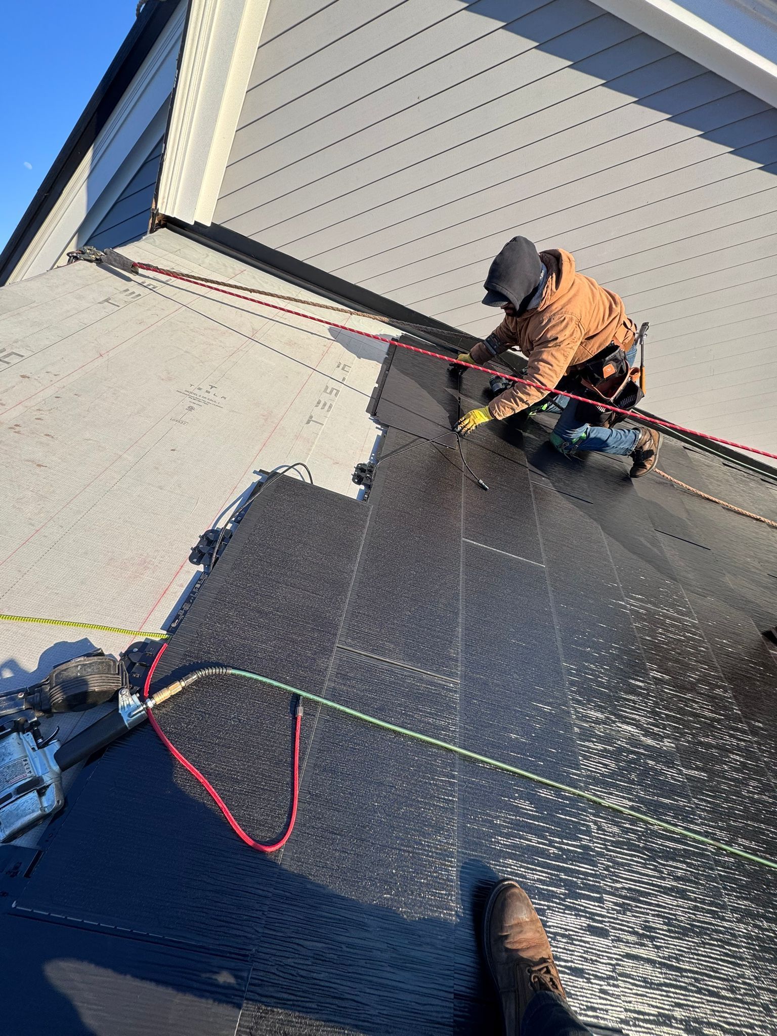 Roofer in safety harness working on a dark roof, connecting cables, near a light-colored building under a blue sky.